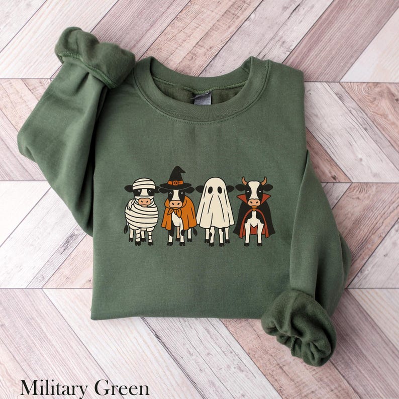 May include: Military green sweatshirt featuring a Halloween-themed design. The graphic depicts four cows dressed as a mummy, witch, ghost, and vampire. The sweatshirt has long sleeves and a crew neck, perfect for the fall season.