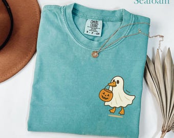 May include: Seafoam green t-shirt with a cartoon ghost duck holding a jack-o'-lantern. The shirt has a Comfort Colors tag and a gold necklace. A brown hat and dried leaves are in the background.