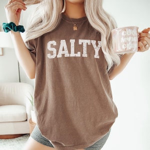 May include: A brown oversized t-shirt with the word "SALTY" printed in white on the front. The shirt is being worn by a person with long blonde hair.