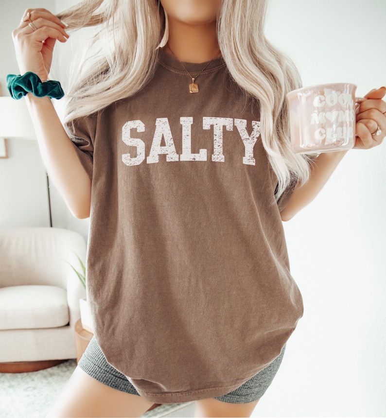 May include: A brown oversized t-shirt with the word "SALTY" printed in white on the front. The shirt is being worn by a person with long blonde hair.