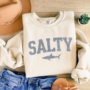 May include: A cream-colored sweatshirt with the word "SALTY" printed in blue with a distressed font. A blue shark silhouette is below the text. The sweatshirt is laid out on a wooden surface with a brown hat, blue jeans, and sandals.