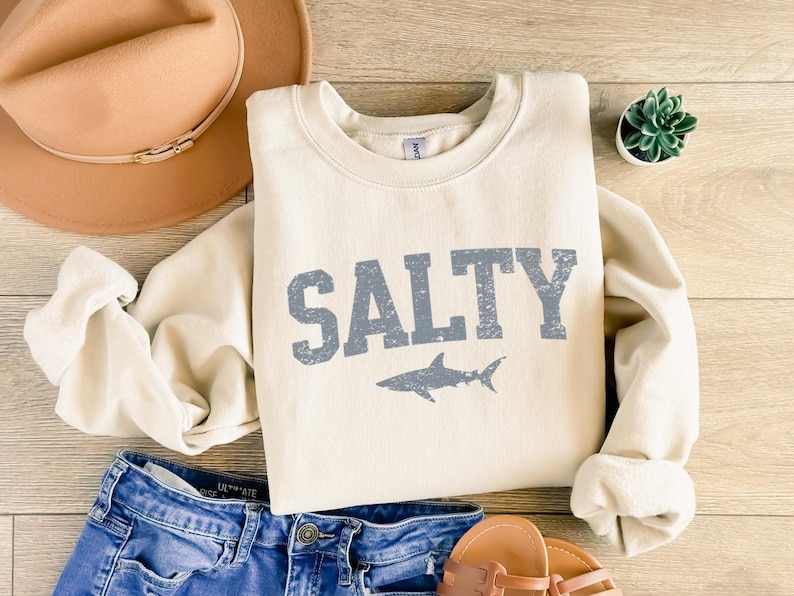 May include: A cream-colored sweatshirt with the word "SALTY" printed in blue with a distressed font. A blue shark silhouette is below the text. The sweatshirt is laid out on a wooden surface with a brown hat, blue jeans, and sandals.