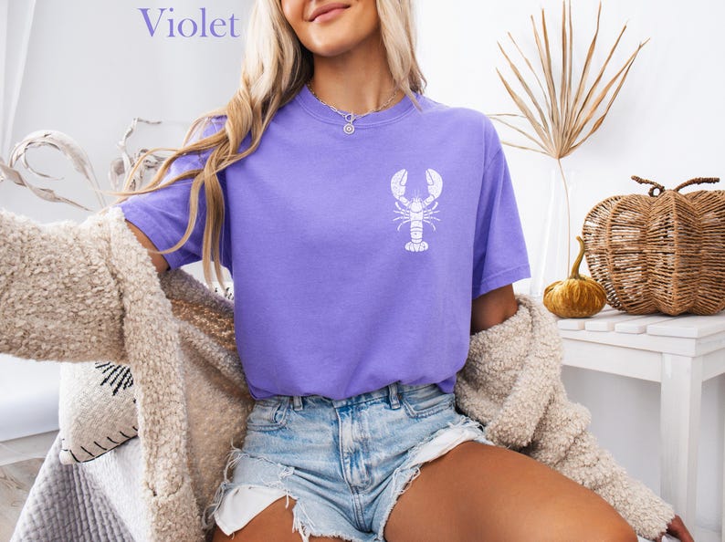 以下が含まれることがあります： 白いロブスターのグラフィックがプリントされたパープルのTシャツを着た女性。Tシャツはゆったりとしたフィット感で、袖は短いです。女性はデニムのショートパンツを履いており、長いブロンドの髪をしています。