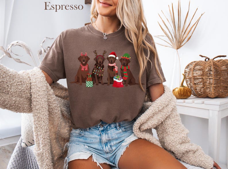 Op de afbeelding: Espresso-kleurig T-shirt met een feestelijk ontwerp van vijf bruine Labrador Retrievers in kerstkleding. Elke hond draagt een ander vakantieaccessoire, waaronder geweien, een kerstmuts en sjaals. Het shirt wordt gecombineerd met een spijkerbroek.