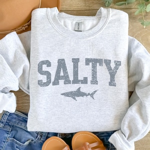 May include: A gray sweatshirt with the word "SALTY" printed in a distressed font in dark gray. A small shark silhouette is printed below the word.