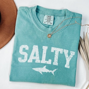 Könnte beinhalten: Ein türkisfarbenes T-Shirt mit dem Wort "SALTY" in Weiß bedruckt, darunter eine weiße Hai-Silhouette. Das Shirt hat einen Used-Look und ist aus einem weichen, bequemen Stoff.