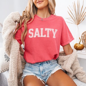 May include: A woman wearing a pink t-shirt with the word "SALTY" printed in white on the front. She is also wearing denim shorts and a white fuzzy cardigan.