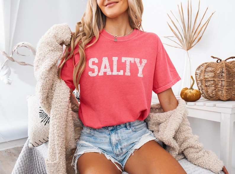 May include: A woman wearing a pink t-shirt with the word "SALTY" printed in white on the front. She is also wearing denim shorts and a white fuzzy cardigan.