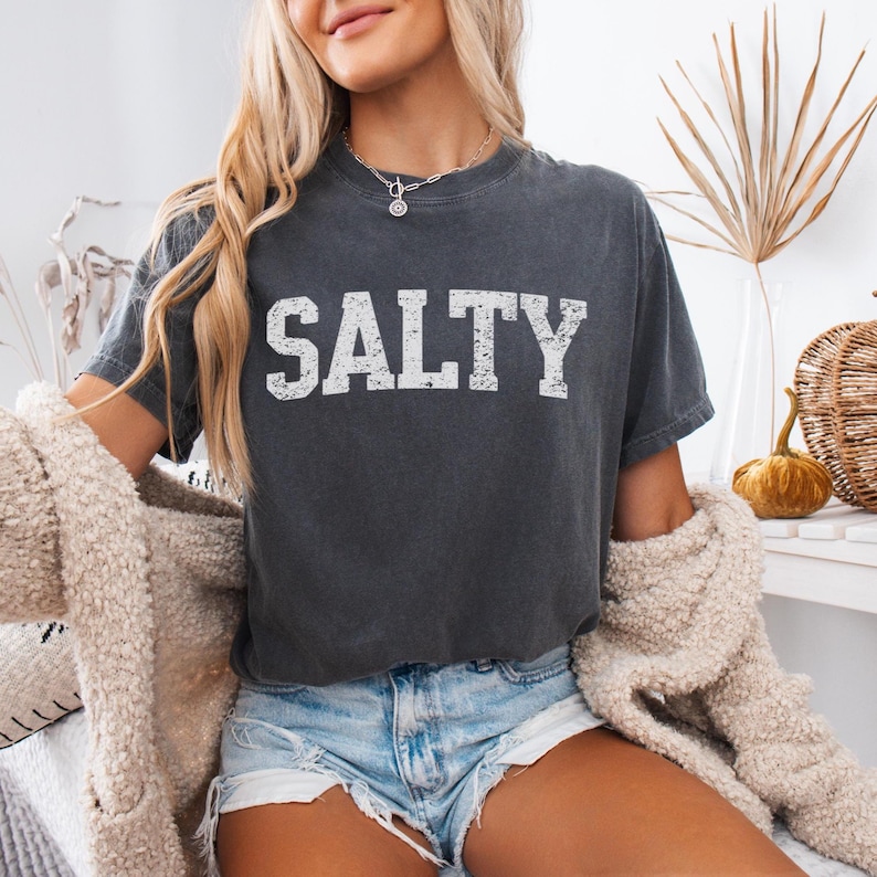 May include: A dark gray t-shirt with the word "SALTY" printed in white on the front. The shirt is worn by a person with long blonde hair and is styled with denim shorts.