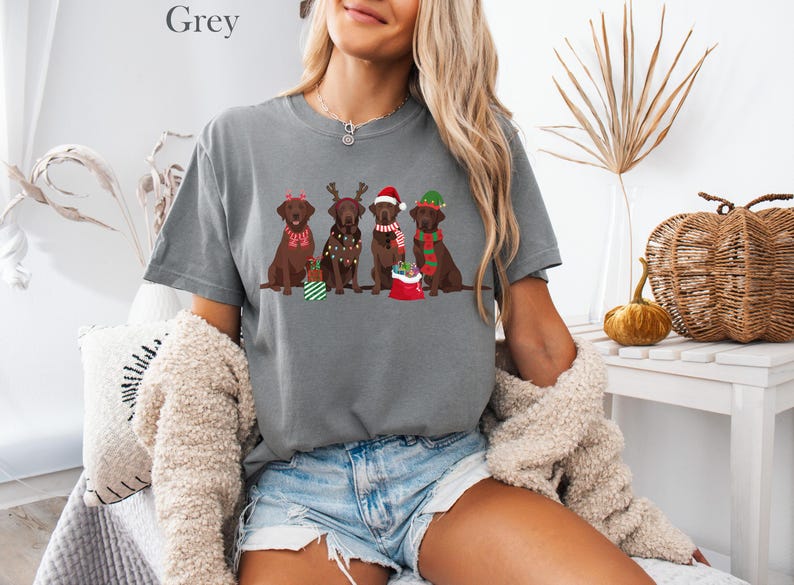 Op de afbeelding: Een grijs T-shirt met een kerstthema. Het toont vier bruine Labradors, elk met feestelijke accessoires zoals kerstmutsen en sjaals. Het woord "Grey" staat bovenaan het T-shirt gedrukt. De honden dragen kerstversieringen.