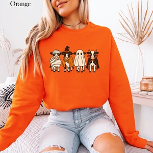 May include: Orange sweatshirt featuring a Halloween-themed graphic of four cows dressed as a mummy, witch, ghost, and vampire. The word "Orange" is printed in the upper left corner. The sweatshirt is a long-sleeved crew neck.