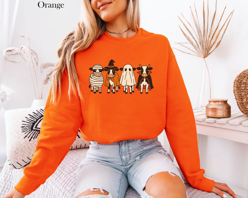 May include: Orange sweatshirt featuring a Halloween-themed graphic of four cows dressed as a mummy, witch, ghost, and vampire. The word "Orange" is printed in the upper left corner. The sweatshirt is a long-sleeved crew neck.