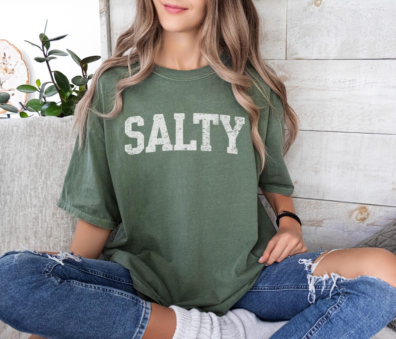 May include: A woman wearing a green t-shirt with the word "SALTY" printed in white on the front. She is sitting on a couch with her legs crossed and wearing blue jeans.