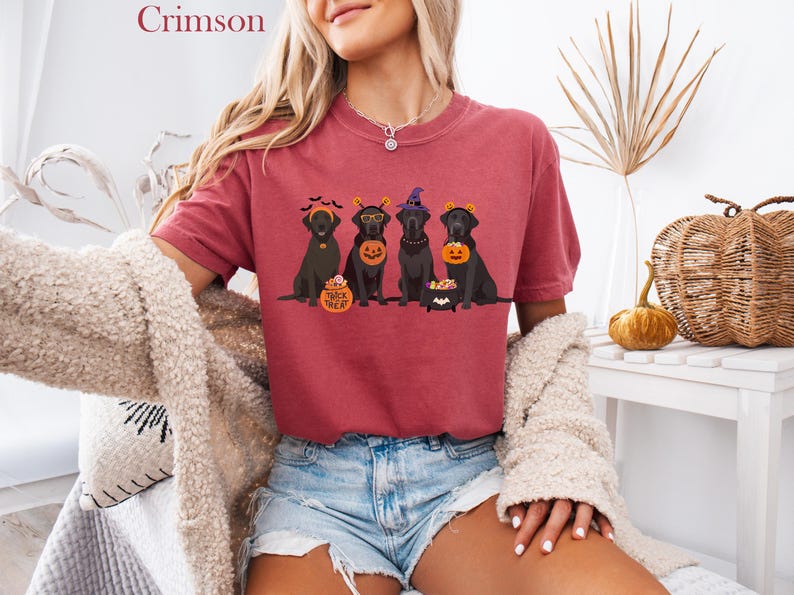 May include: Crimson-colored t-shirt featuring a Halloween-themed graphic of four black Labrador Retrievers. Each dog is dressed in a costume, holding a pumpkin or a cauldron. The shirt is paired with denim shorts.