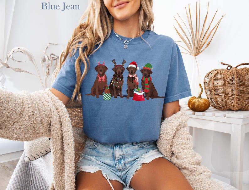 Op de afbeelding: Blauw spijkerbroekkleurig T-shirt met vier bruine Labrador Retrievers met feestelijke vakantieaccessoires. Elke hond heeft een unieke kerstthema-outfit, waaronder kerstmutsen, sjaals en rendiergeweien. Op de shirt staat bovenaan "Blue Jean".