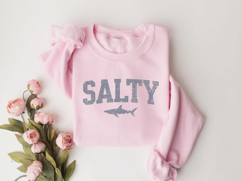 May include: Pink sweatshirt with the word "SALTY" in gray text and a gray shark silhouette below it.