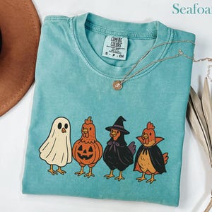 May include: Seafoam green t-shirt with a graphic of four chickens in Halloween costumes: a ghost, a pumpkin, a witch, and a vampire. The shirt has a crew neck and short sleeves. A gold necklace is draped across the shirt.