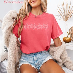 May include: A woman wearing a pink t-shirt with a white graphic of four sea turtles. The shirt is a relaxed fit and has a distressed look. The woman is also wearing denim shorts and a white fuzzy cardigan.