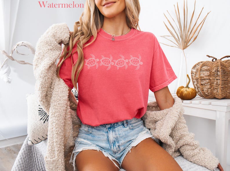 May include: A woman wearing a pink t-shirt with a white graphic of four sea turtles. The shirt is a relaxed fit and has a distressed look. The woman is also wearing denim shorts and a white fuzzy cardigan.