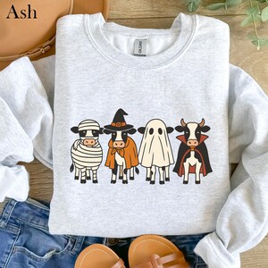 May include: Ash-colored sweatshirt featuring a Halloween-themed design. The design includes four cartoon cows dressed as a mummy, witch, ghost, and vampire. The sweatshirt is displayed on a wooden surface with a pair of sandals and jeans.