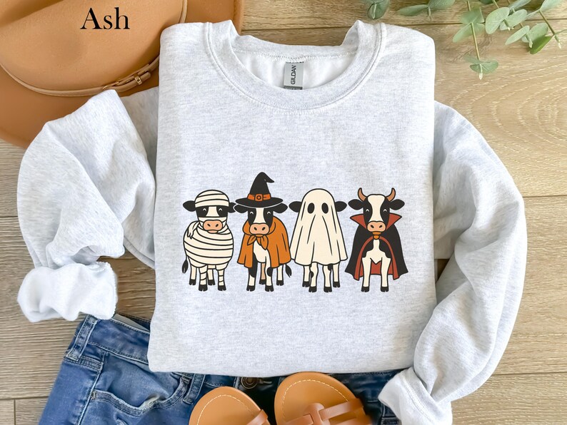 May include: Ash-colored sweatshirt featuring a Halloween-themed design. The design includes four cartoon cows dressed as a mummy, witch, ghost, and vampire. The sweatshirt is displayed on a wooden surface with a pair of sandals and jeans.