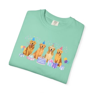 May include: A light green t-shirt featuring a graphic of four golden retriever dogs wearing party hats. The dogs are sitting near a birthday cake and wrapped gifts. The words "Happy Birthday" are on the cake.