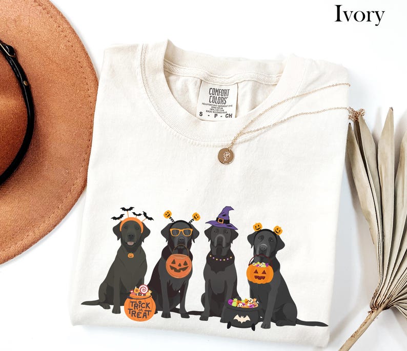 May include: Ivory-colored t-shirt featuring four black Labrador Retrievers dressed in Halloween costumes. The dogs wear witch hats, pumpkin headbands, and carry trick-or-treat buckets. The shirt has the text "Trick or Treat".