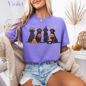 May include: A violet-colored t-shirt featuring a Halloween-themed graphic of four black Labrador Retrievers. Each dog is dressed in a costume, holding a pumpkin or a treat bucket. The word "Violet" is printed at the top.
