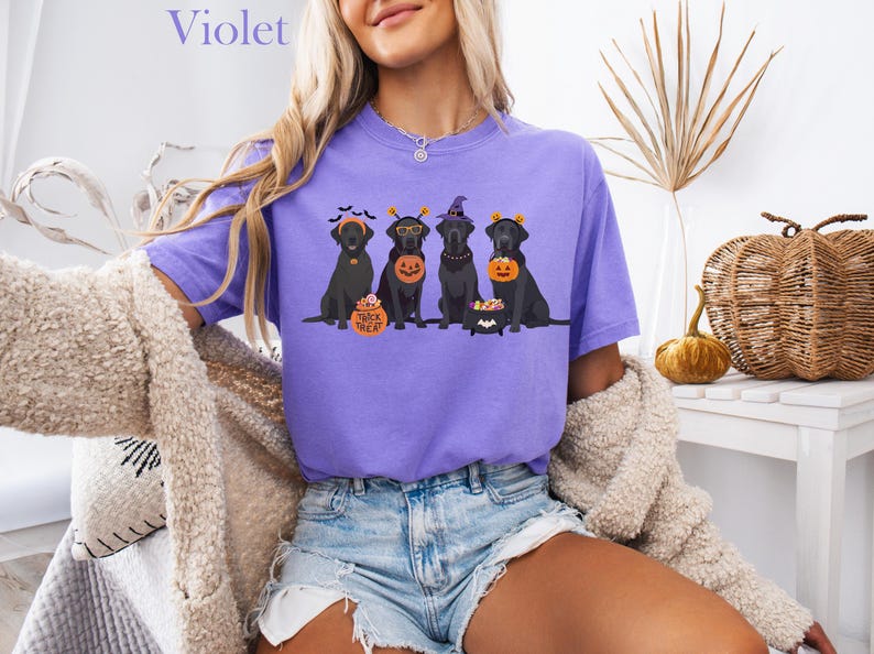 May include: A violet-colored t-shirt featuring a Halloween-themed graphic of four black Labrador Retrievers. Each dog is dressed in a costume, holding a pumpkin or a treat bucket. The word "Violet" is printed at the top.
