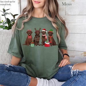 Op de afbeelding: Mosgroen t-shirt met een afbeelding van vijf bruine Labradors in kerstoutfits. De honden dragen feestelijke accessoires zoals rendiergeweien, kerstmutsen en sjaals. De tekst "Model has sized up for the oversized look" is zichtbaar.