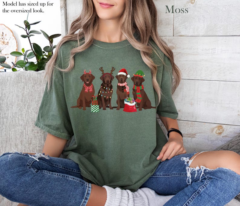 Op de afbeelding: Mosgroen t-shirt met een afbeelding van vijf bruine Labradors in kerstoutfits. De honden dragen feestelijke accessoires zoals rendiergeweien, kerstmutsen en sjaals. De tekst "Model has sized up for the oversized look" is zichtbaar.