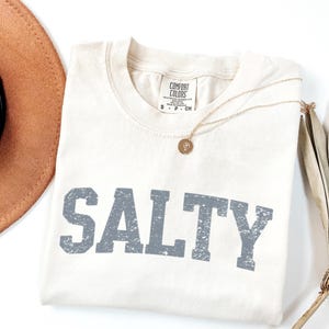 May include: A cream colored t-shirt with the word "SALTY" printed in gray on the front. The shirt has a distressed, vintage look.