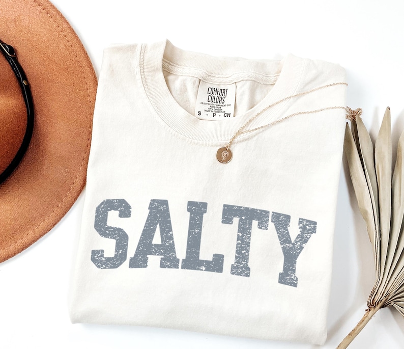 May include: A cream colored t-shirt with the word "SALTY" printed in gray on the front. The shirt has a distressed, vintage look.