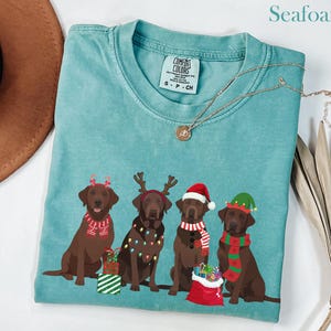 Könnte beinhalten: Türkisfarbenes T-Shirt mit einem Aufdruck von vier braunen Labrador-Retrievern in festlicher Kleidung. Einer trägt ein Rentiergeweih, ein anderer einen Weihnachtsmannhut und ein weiterer einen Elfenhut. Das Shirt hat ein Comfort Colors-Etikett.