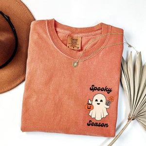 May include: A coral colored t-shirt with a vintage-style graphic of a ghost holding a pumpkin and a flower. The text "Spooky Season" is printed below the ghost.