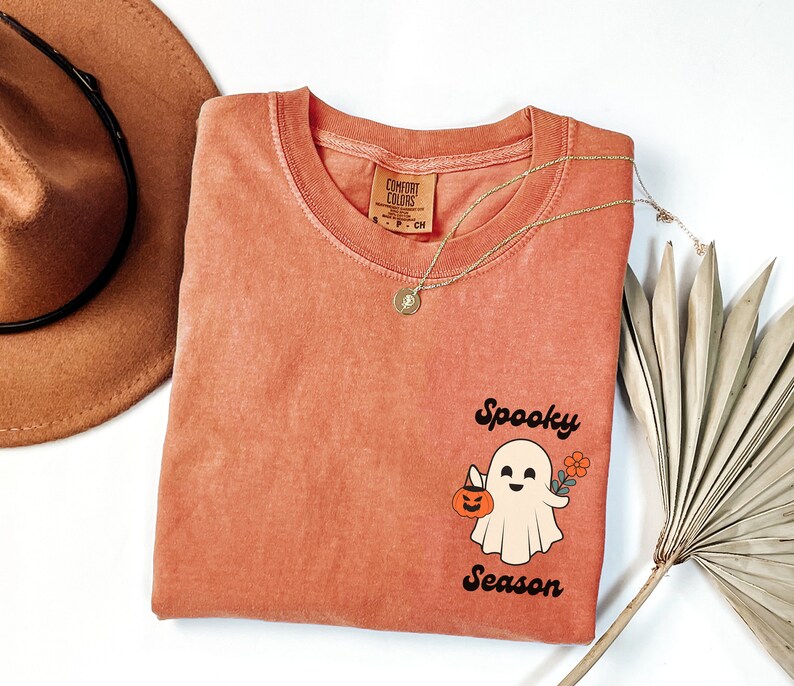 May include: A coral colored t-shirt with a vintage-style graphic of a ghost holding a pumpkin and a flower. The text "Spooky Season" is printed below the ghost.
