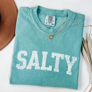 May include: A teal blue t-shirt with the word "SALTY" printed in white distressed lettering. The shirt is made of a soft, comfortable fabric and has a relaxed fit.