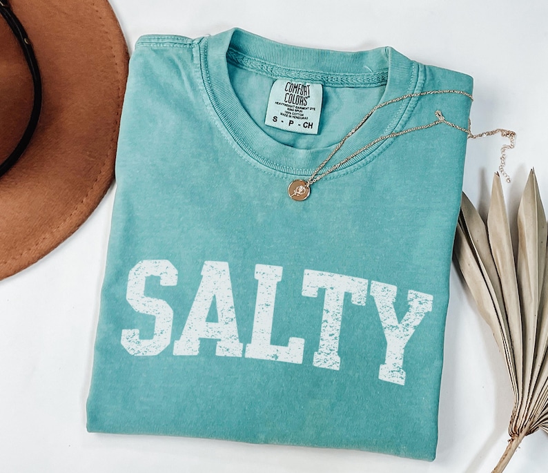 May include: A teal blue t-shirt with the word "SALTY" printed in white distressed lettering. The shirt is made of a soft, comfortable fabric and has a relaxed fit.