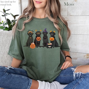 May include: Moss green t-shirt featuring a Halloween-themed graphic of five black Labrador Retrievers wearing festive costumes. The dogs are adorned with witch hats, pumpkin buckets, and trick-or-treat accessories. The text "Trick or Treat" is on a pumpkin.