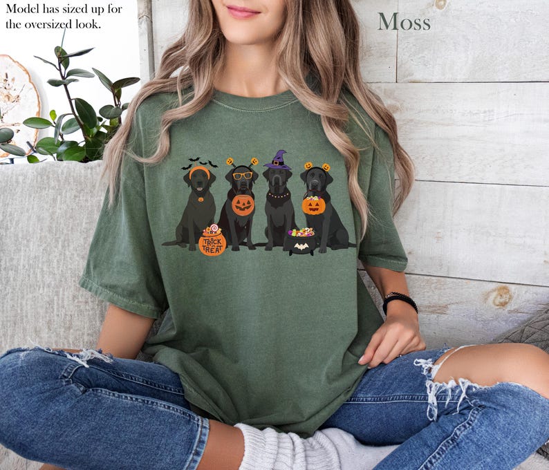 May include: Moss green t-shirt featuring a Halloween-themed graphic of five black Labrador Retrievers wearing festive costumes. The dogs are adorned with witch hats, pumpkin buckets, and trick-or-treat accessories. The text "Trick or Treat" is on a pumpkin.