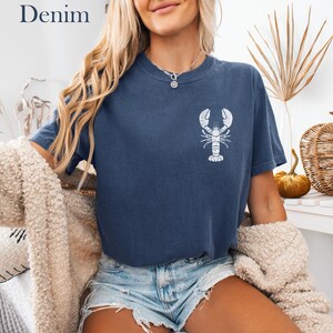 以下が含まれることがあります： 白いロブスターのグラフィックがプリントされたネイビーのTシャツを着た女性。デニムのショートパンツと白いふわふわのカーディガンを着ています。