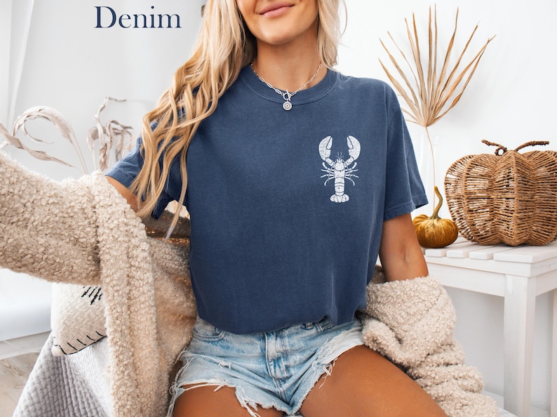 以下が含まれることがあります： 白いロブスターのグラフィックがプリントされたネイビーのTシャツを着た女性。デニムのショートパンツと白いふわふわのカーディガンを着ています。