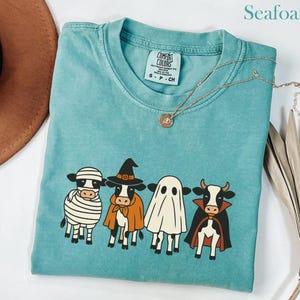 Puede incluir: Camiseta verde mar con un gráfico de cuatro vacas disfrazadas para Halloween: una momia, una bruja, un fantasma y un vampiro. La camiseta tiene cuello redondo y una etiqueta Comfort Colors. Un collar dorado está drapeado sobre la camiseta.