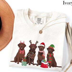 Op de afbeelding: Ivoor kleurig T-shirt met vier bruine Labrador Retrievers in feestelijke kerstkleding. Elke hond draagt een ander accessoire, waaronder een kerstmuts, een elfenmuts en rendiergeweien. Het shirt heeft ook de tekst "COMFORT COLORS".