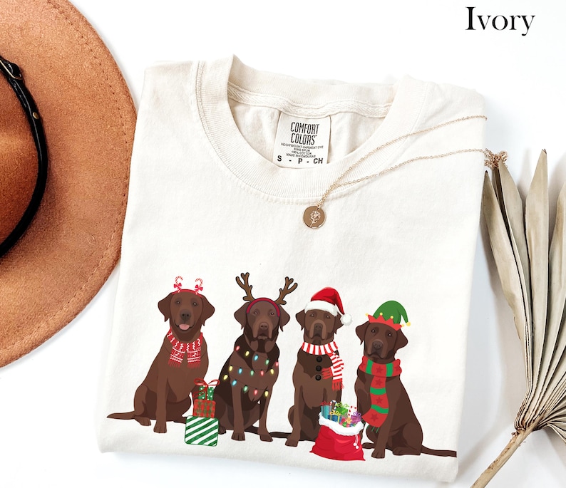 Op de afbeelding: Ivoor kleurig T-shirt met vier bruine Labrador Retrievers in feestelijke kerstkleding. Elke hond draagt een ander accessoire, waaronder een kerstmuts, een elfenmuts en rendiergeweien. Het shirt heeft ook de tekst "COMFORT COLORS".