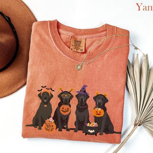 May include: A yam-colored Comfort Colors t-shirt featuring a Halloween-themed graphic. The graphic depicts four black Labrador Retrievers in Halloween costumes, including witch hats and pumpkin buckets. The words "Trick Treat" are on a candy bucket.