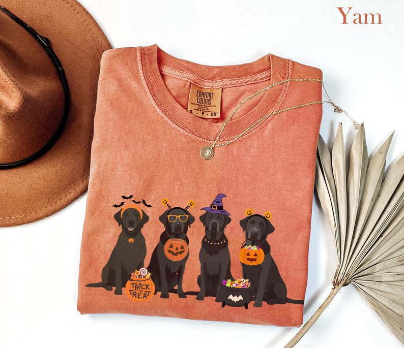 May include: A yam-colored Comfort Colors t-shirt featuring a Halloween-themed graphic. The graphic depicts four black Labrador Retrievers in Halloween costumes, including witch hats and pumpkin buckets. The words "Trick Treat" are on a candy bucket.