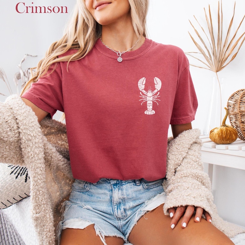 以下が含まれることがあります： 白いロブスターのグラフィックがプリントされた赤いTシャツを着た女性。Tシャツはクルーネックで半袖。女性はデニムのショートパンツと白いカーディガンを着ています。