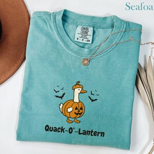 May include: Seafoam green t-shirt with a pumpkin-themed duck graphic and the text "Quack-O'-Lantern." The shirt has a crew neck and short sleeves. A gold necklace with a pendant is draped across the shirt.