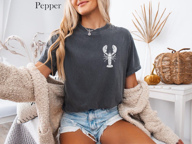 以下が含まれることがあります： 白いロブスターのグラフィックがプリントされたダークグレーのTシャツ。ゆったりとしたフィット感で、クルーネックです。